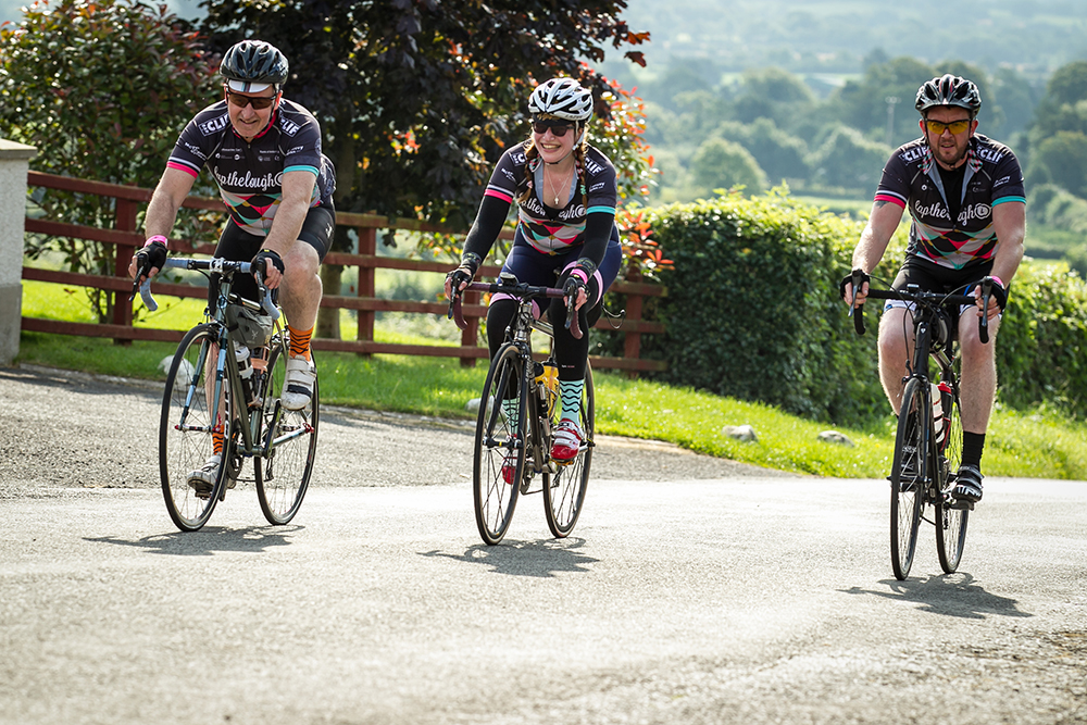 Three cyclists smile and laugh together as they ride up a country road on a sunny summer day