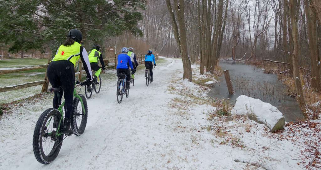 A group of brightly clad cyclist ride up a snowy trail by a wooden fence and a frozen pond
