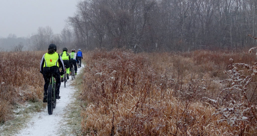 Four cyclists ride away on a lightly snow covered train in a brown grassy field