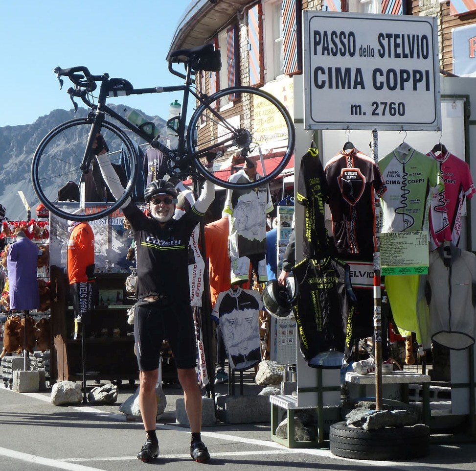 A white bearded cyclist proudly holds his bicycle up over his head next to a sign that reads 'Passo Dello Stelvio Cima Coppi m.2760'