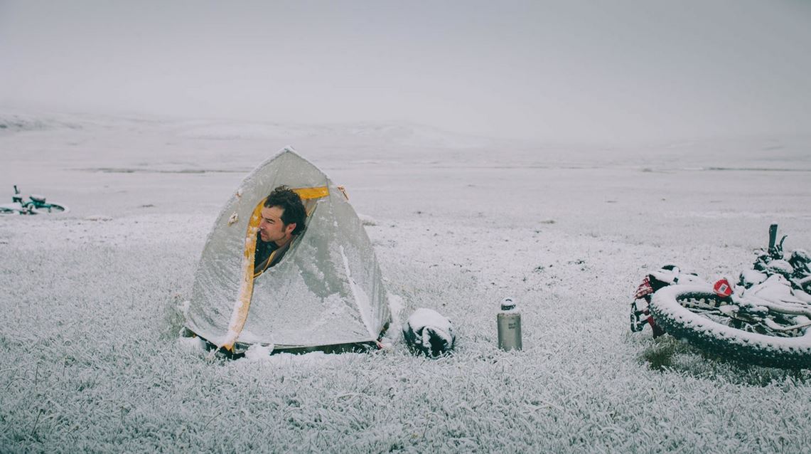 A man's head pokes out of a bivy sack to face a raw winter wind. Snow covers everthing, including his bicycle which rests on the ground next to him.