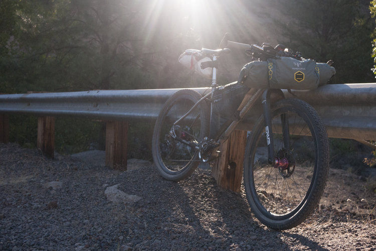 A Seven loaded for bikepacking leans against a guardrial, the sun cascading in from behind