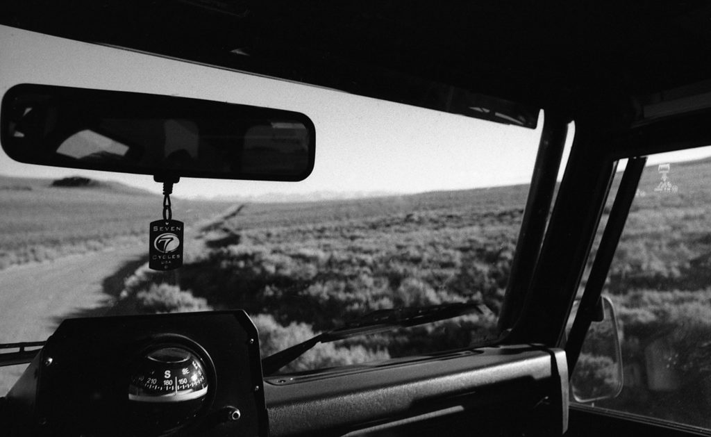 the silhouette of a Seven head badge key chain hangs on the rear view mirror of a truck driving down a lonely road