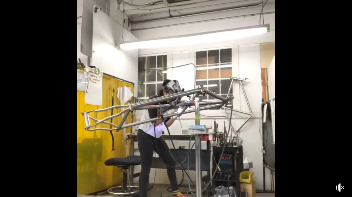 a welder welding a titanium tandem on her workstand in front of her bench