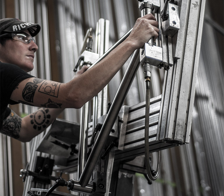 low angle photo of a machininst setting up a titanium bicycle frame in her jig, in front of a wall of titanium tubes